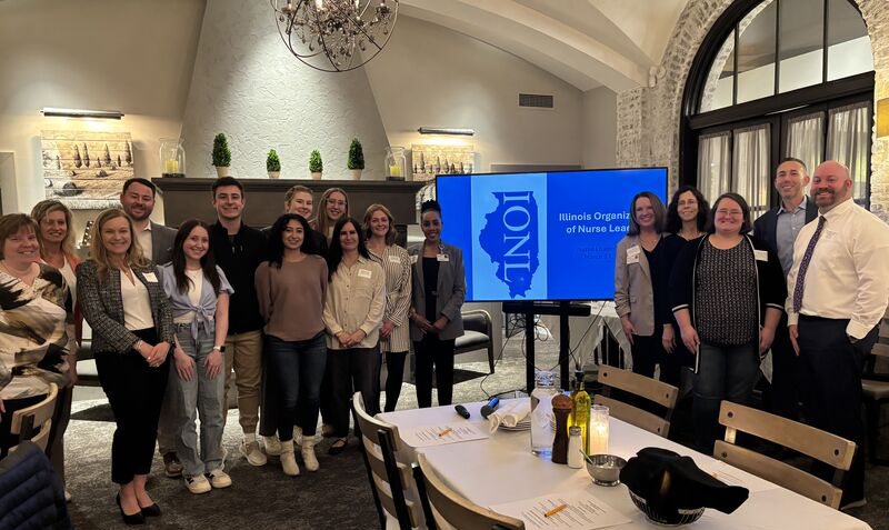 A group of people stand in front of a screen displaying the Illinois Organization of Nurse Leaders logo. The group appears to be attending a conference or event.