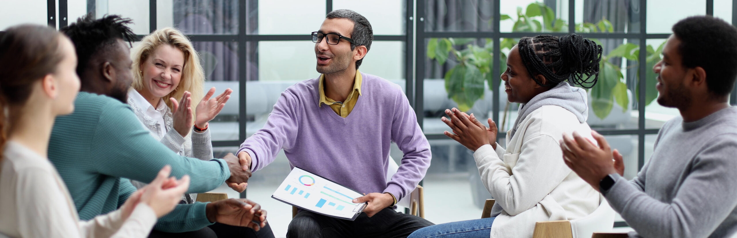 A group of people sitting in a circle, with two individuals shaking hands in the center while others clap and smile. One person holds a document with charts and graphs.