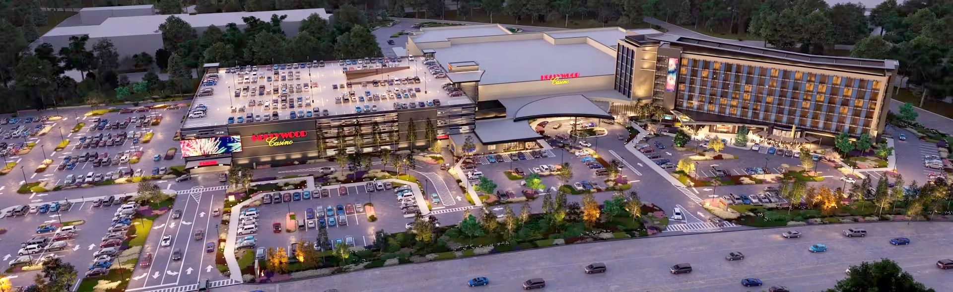Aerial view of the Hollywood Casino Aurora with a multi-story parking garage and many cars in the surrounding lots. The buildings are lit up, and trees surround the property.